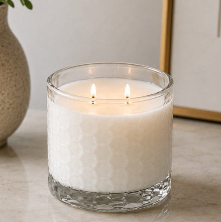 A lit white candle in a clear textured glass jar sits on a light marble surface, with a speckled beige vase of greenery and part of a framed artwork in the background.