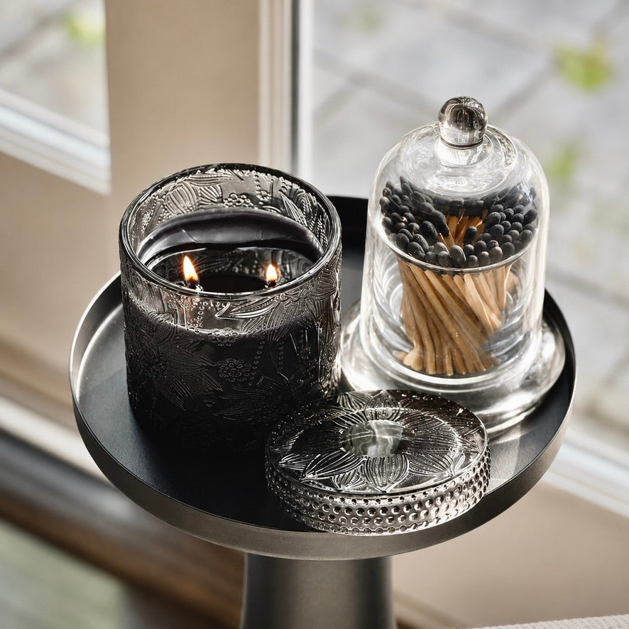 A black textured glass candle jar with two lit wicks sits on a round black side table next to a clear glass cloche holding wooden matches, with a matching engraved glass lid in front. The setup is photographed indoors near a window.