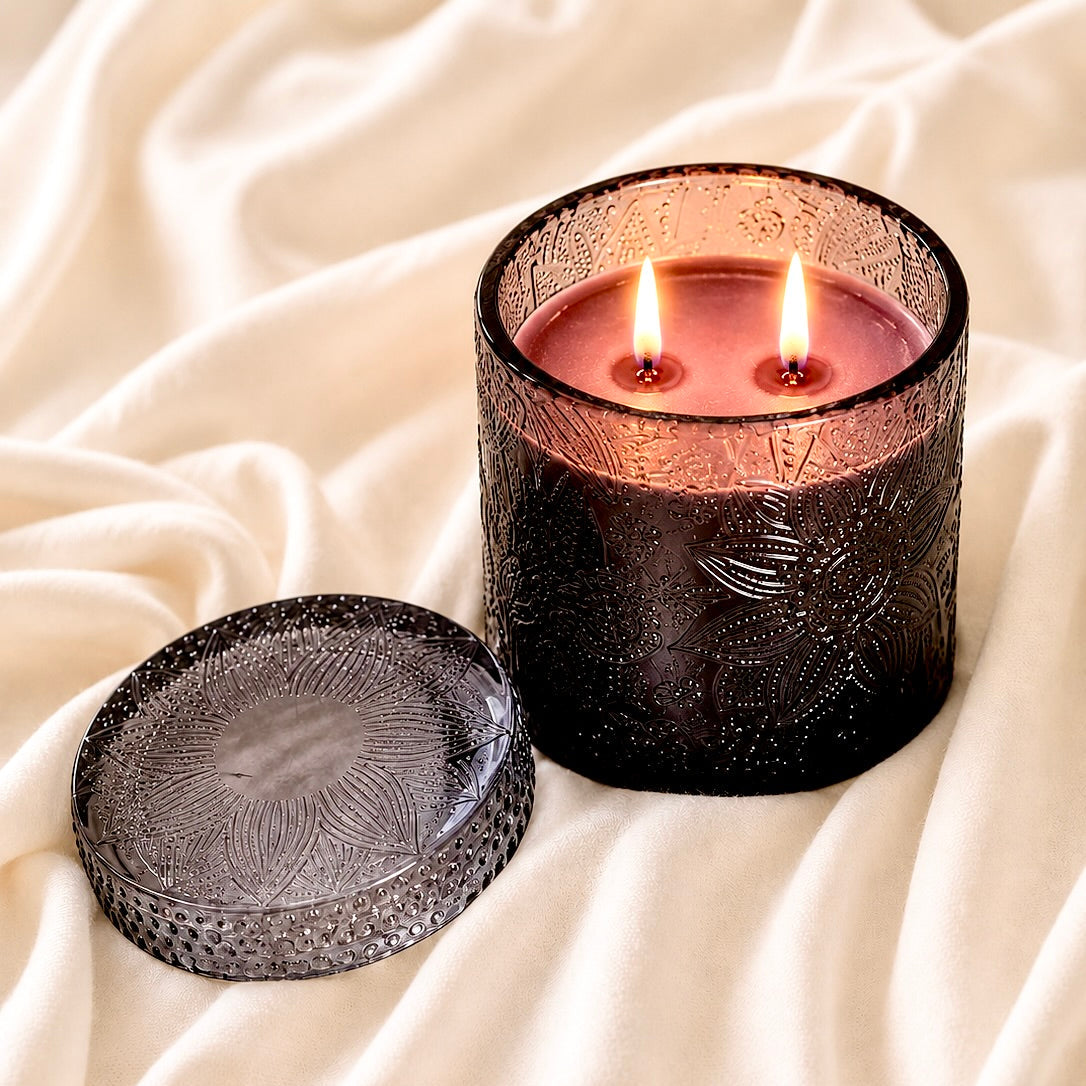 A lit double-wick candle in a dark textured glass holder sits on draped cream fabric, with the matching glass lid placed beside it.