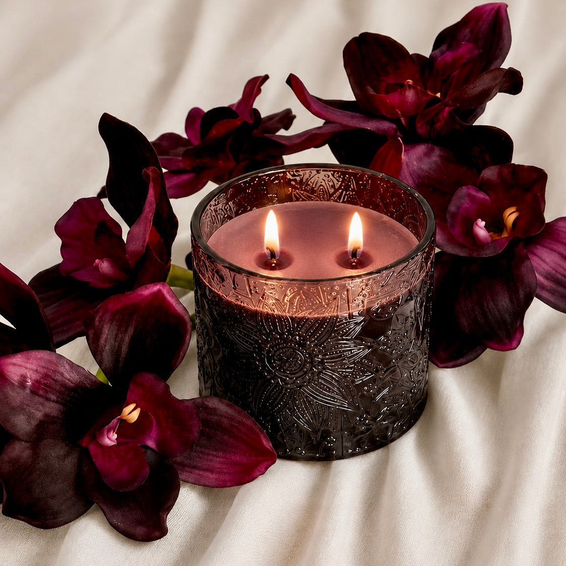 A lit double-wick candle in an embossed dark glass jar, surrounded by deep burgundy orchids on a cream-colored fabric background.