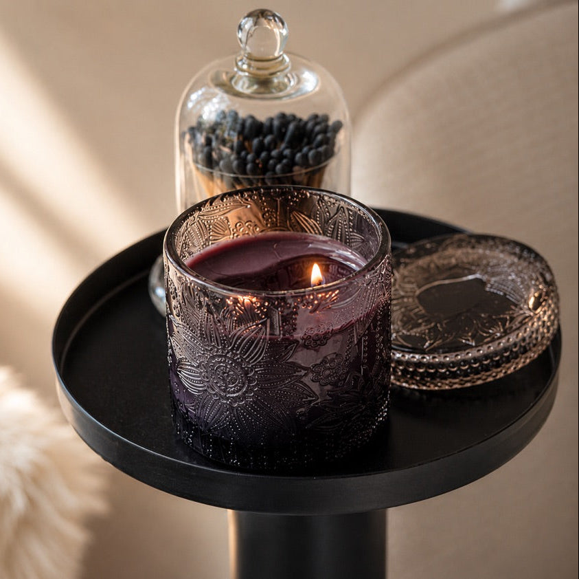 A lit candle in an etched black glass jar sits on a black side table, with a matching decorative lid beside it and a glass cloche holding matches in the background.