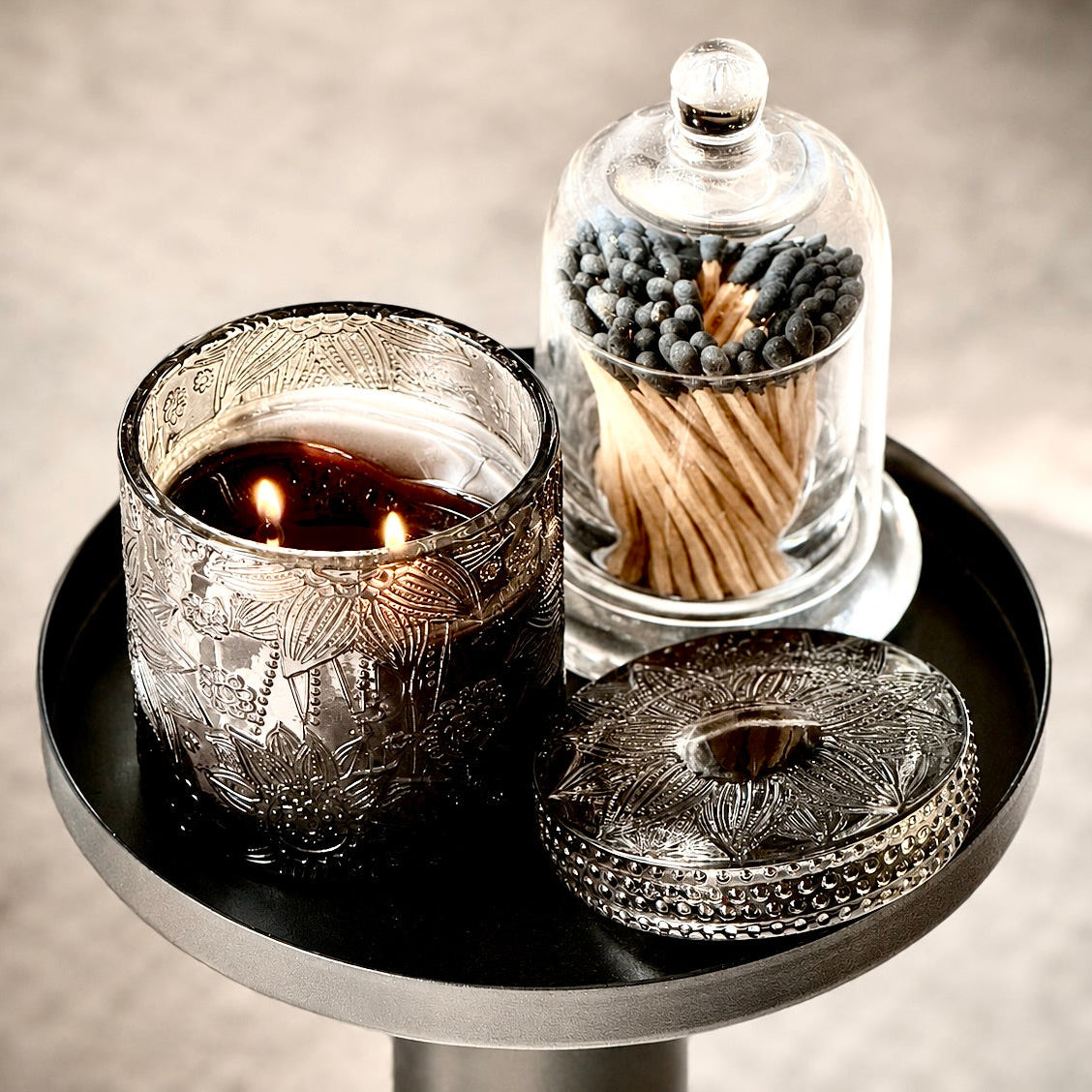 An etched silver glass candle holder with two lit candles sits on a round black tray next to a glass cloche of wooden matches and a matching silver lid.