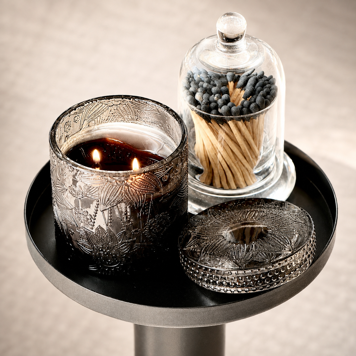 A lit double-wick candle in an etched dark glass holder sits on a round black tray, alongside a glass cloche holding matches and a matching etched lid, against a softly lit neutral background.