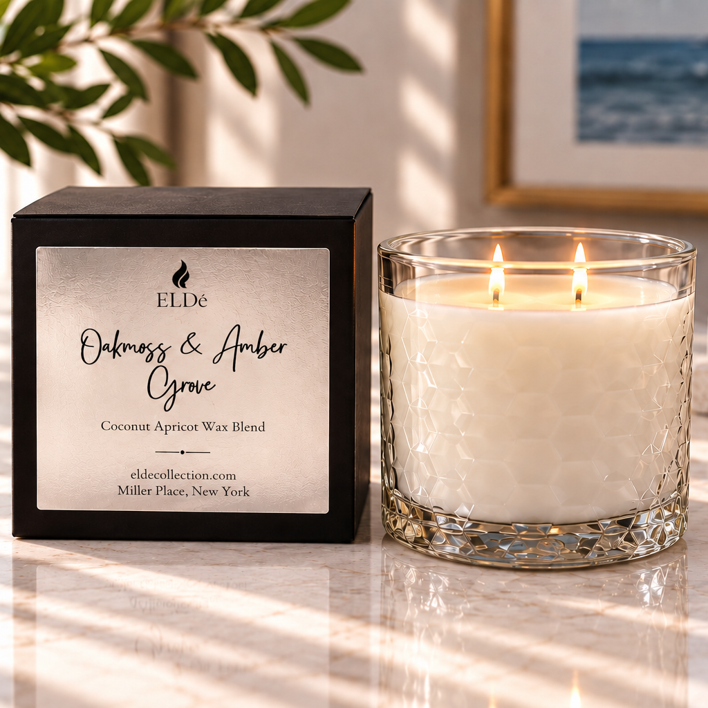 A lit double-wick scented candle in a textured clear glass jar sits beside its black rectangular box on a sunlit marble surface. The box label reads ELDE and 'Olfactory & Amber Grove Coconut Apricot Wax Blend.