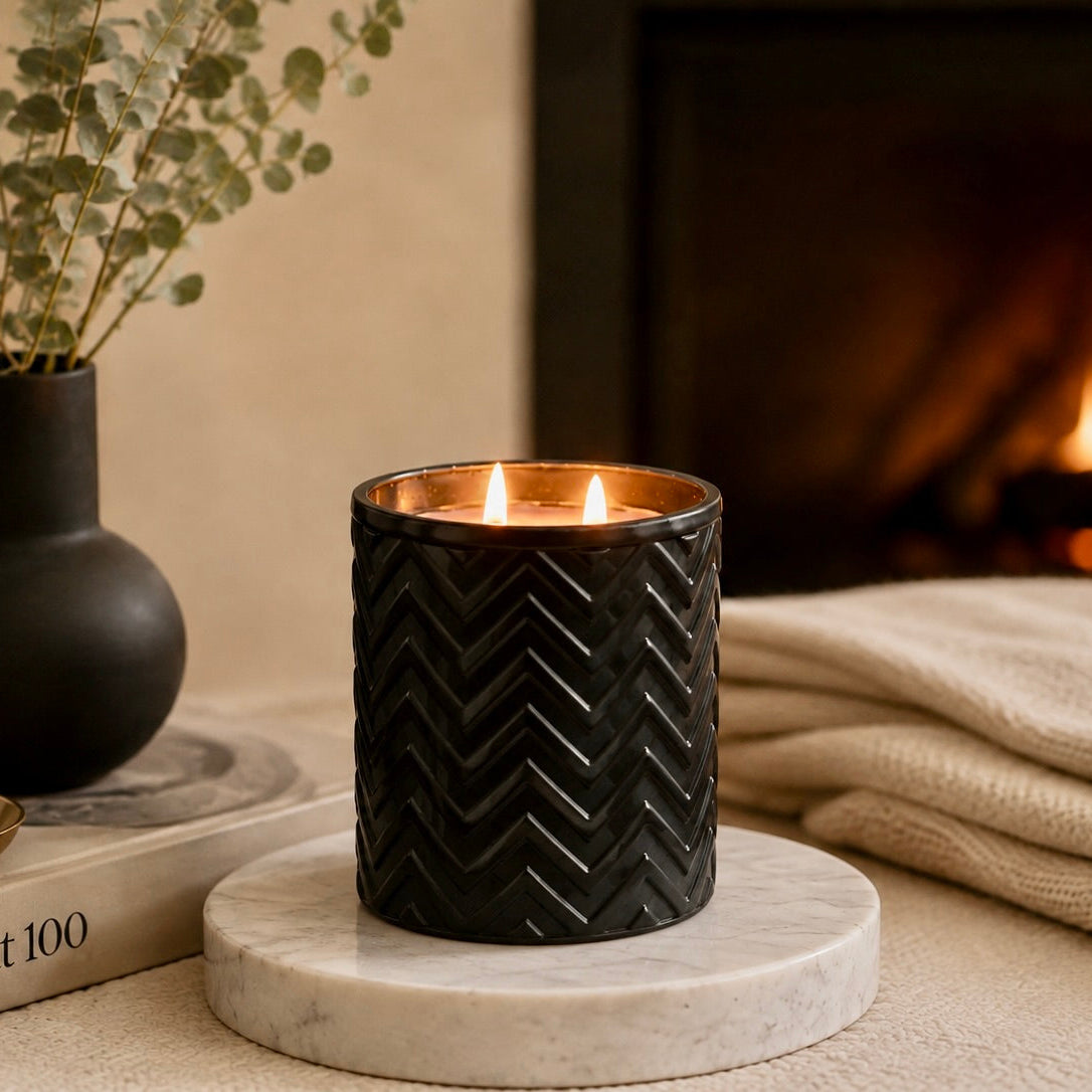 A black textured candle holder with two lit candles sits on a round white marble coaster in a cozy indoor setting. A dark vase with dried leaves, a book, and a folded cream blanket are visible in the background near a blurred fireplace.