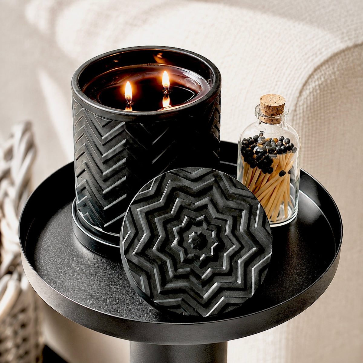 A black textured candle jar with two lit wicks sits on a round black tray beside a small glass bottle of matches, against a soft neutral fabric background.