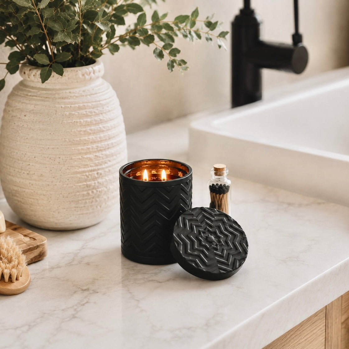 A black textured candle holder with a lit candle and its matching lid on a white marble bathroom counter, styled with a large textured cream vase, soap, wooden brush, and a small bottle.
