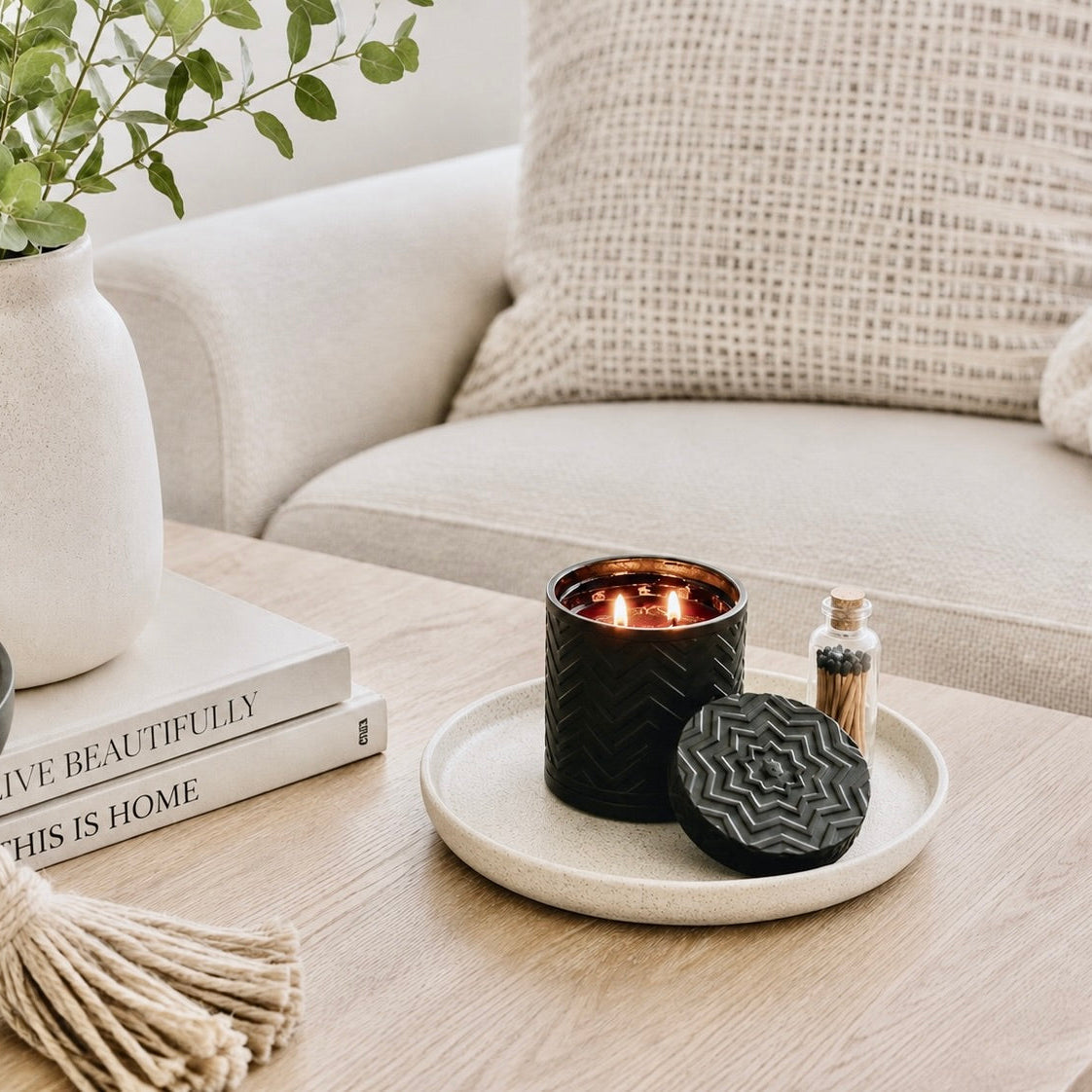 A black textured candle holder with a lit candle and matching lid on a ceramic tray, styled on a wooden table next to books and a vase in a cozy living room setting.
