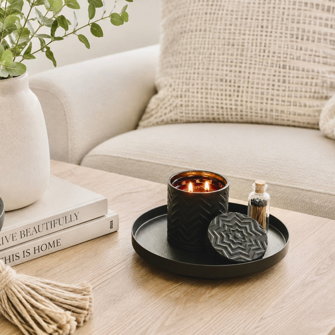 A black textured candle holder with a lit candle and matching lid on a wooden coffee table, styled in a cozy living room setting with a sofa, books, and a plant.