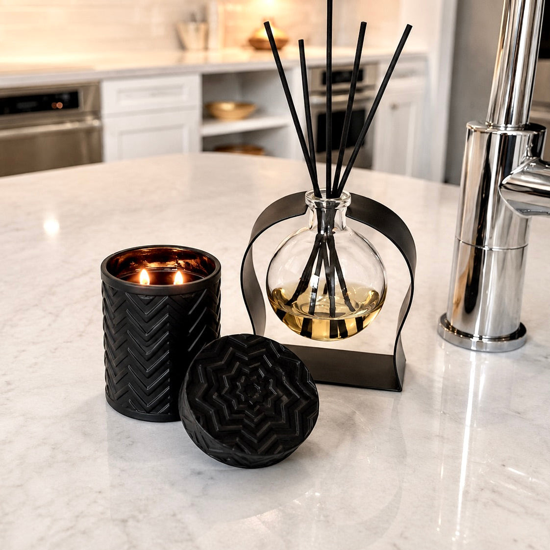 A black ceramic candle holder with a lit candle and a matching round textured lid sit on a white marble kitchen island next to a reed diffuser with black reeds in a glass bottle.