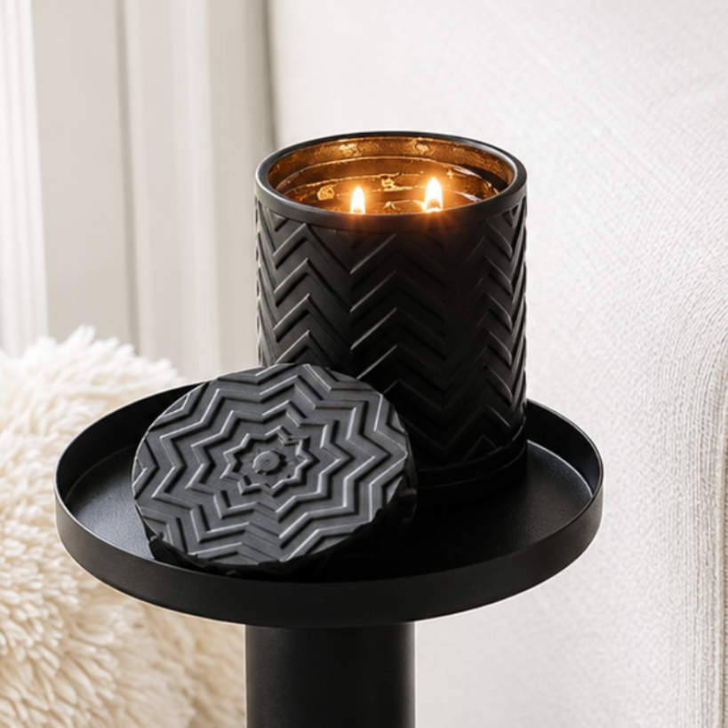 A black textured candle holder with a matching lid sits on a black side table next to a light beige sofa in a bright, modern living room.