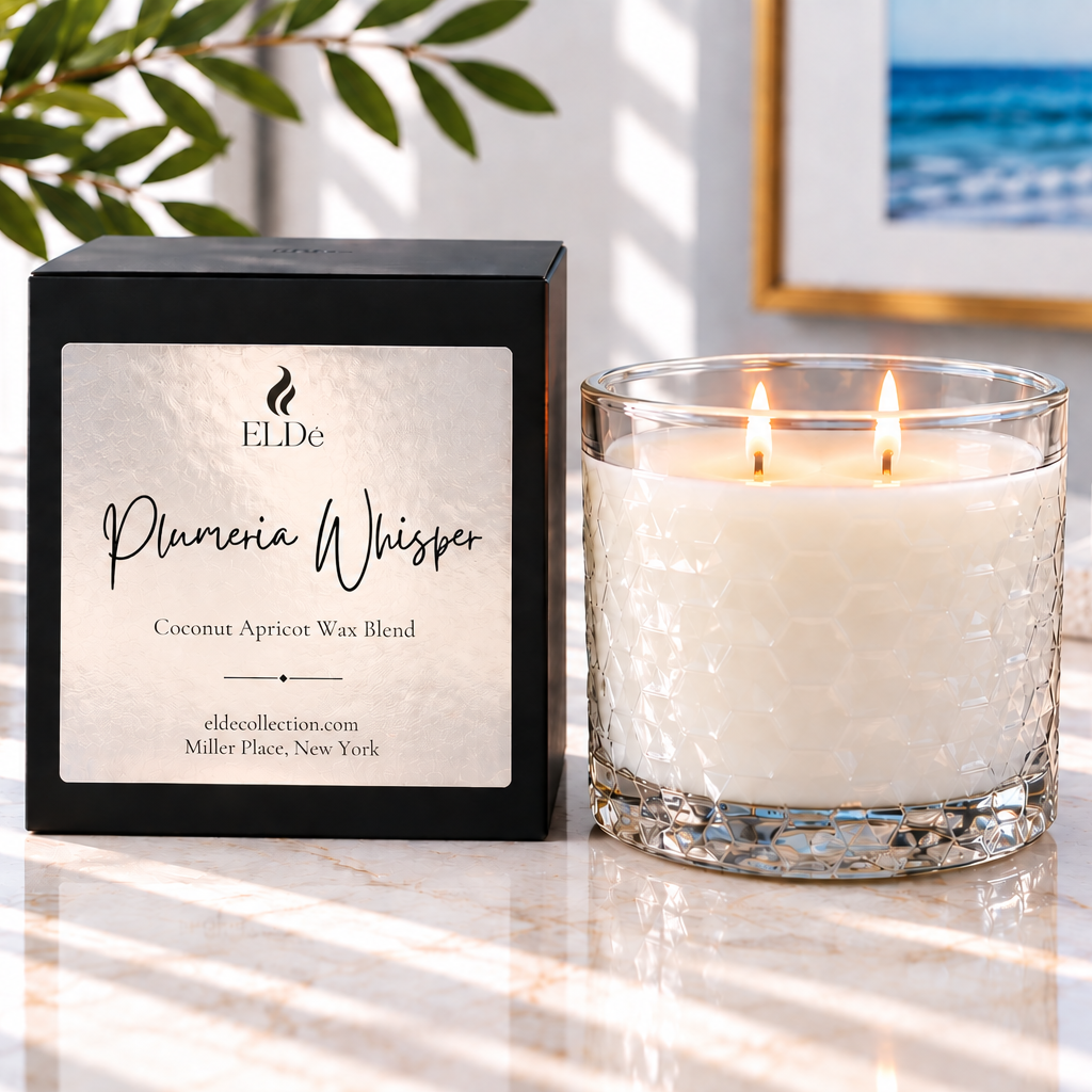 ELDe 'Plumeria Whisper' coconut apricot wax blend candle in a clear textured glass jar with two lit wicks, shown next to its black packaging box on a marble surface with natural sunlight and shadows.