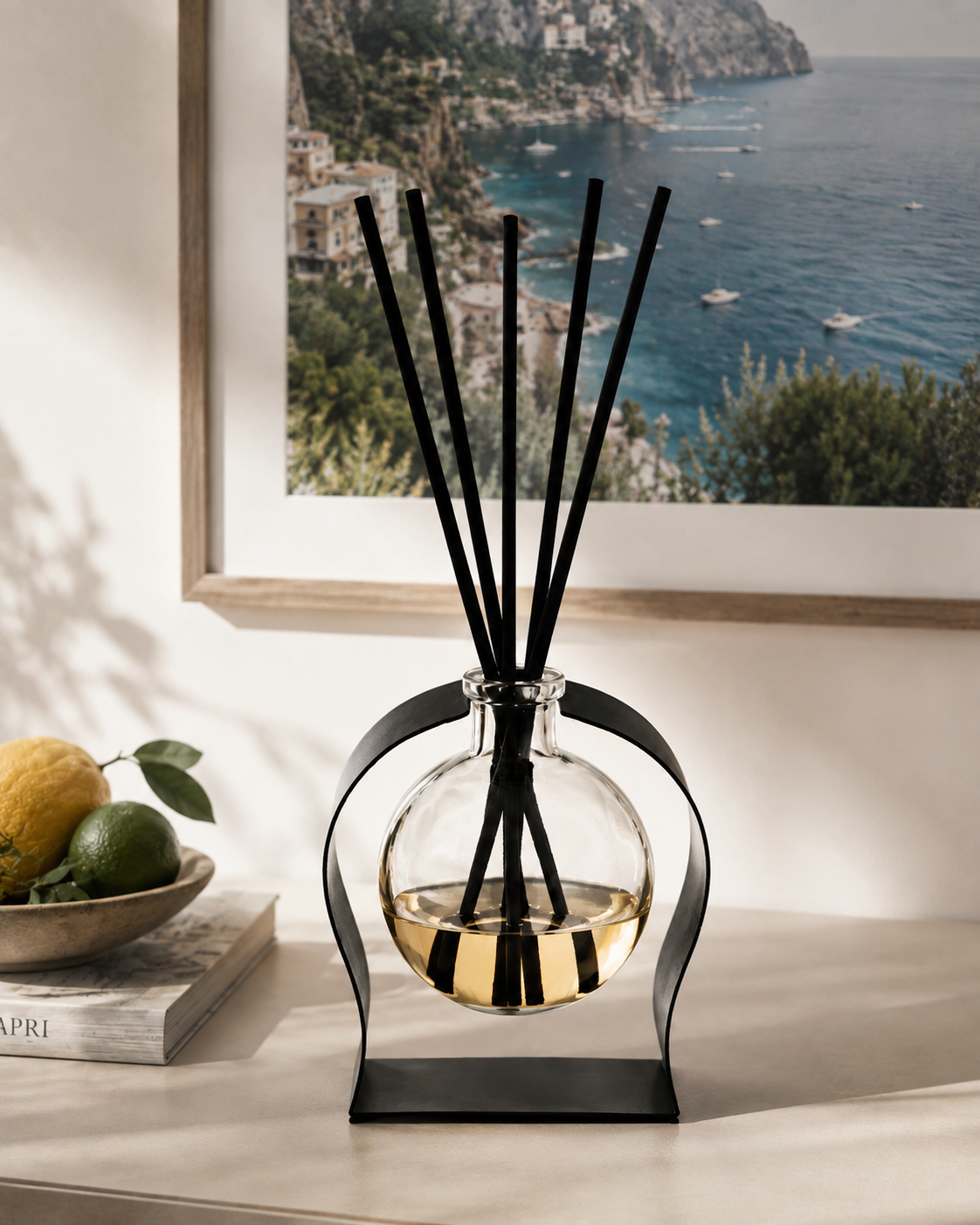 A reed diffuser with a clear glass round bottle, black metal stand and reeds, filled with amber liquid, displayed on a tabletop in a styled interior setting with a framed coastal print and lemons in the background.