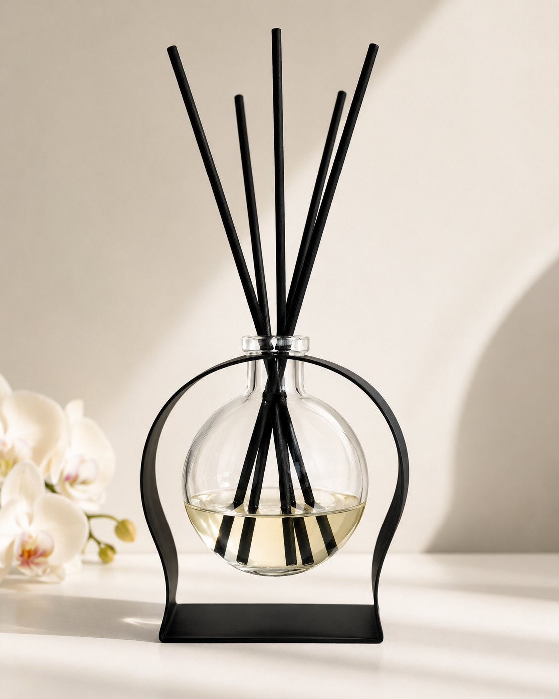 A reed diffuser with a clear glass round bottle, pale yellow liquid, and multiple black reeds, displayed on a black metal stand against a warm beige background with soft shadows and white orchids at the side.