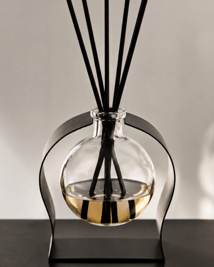 A modern reed diffuser with a clear glass round bottle containing a golden liquid, supported by a curved black metal stand. Five black reed sticks are inserted into the neck of the bottle, set against a neutral light gray background with soft shadows.