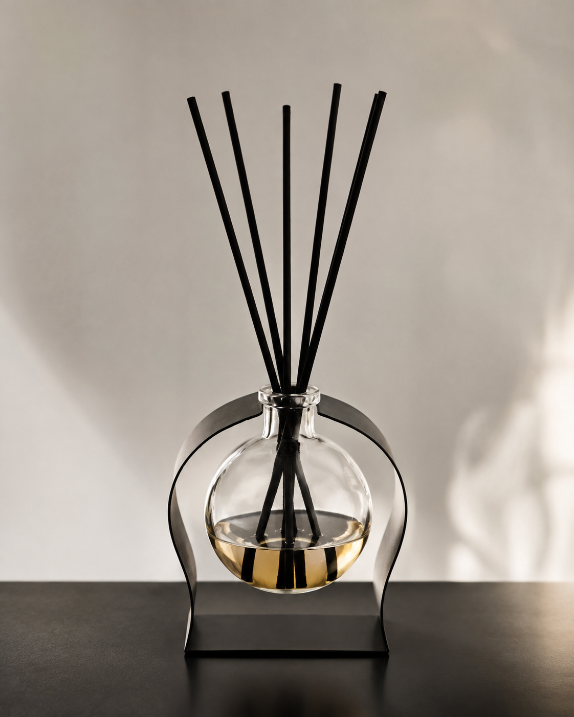 A modern reed diffuser with a clear glass round bottle containing a golden liquid, supported by a curved black metal stand. Five black reed sticks are inserted into the neck of the bottle, set against a neutral light gray background with soft shadows.