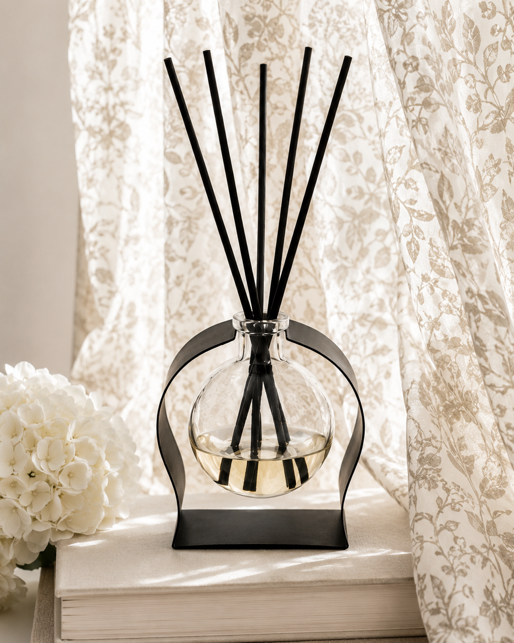 A reed diffuser with a clear glass bottle, black liquid, and black reeds, displayed on a stack of books next to white flowers in front of a beige damask curtain.
