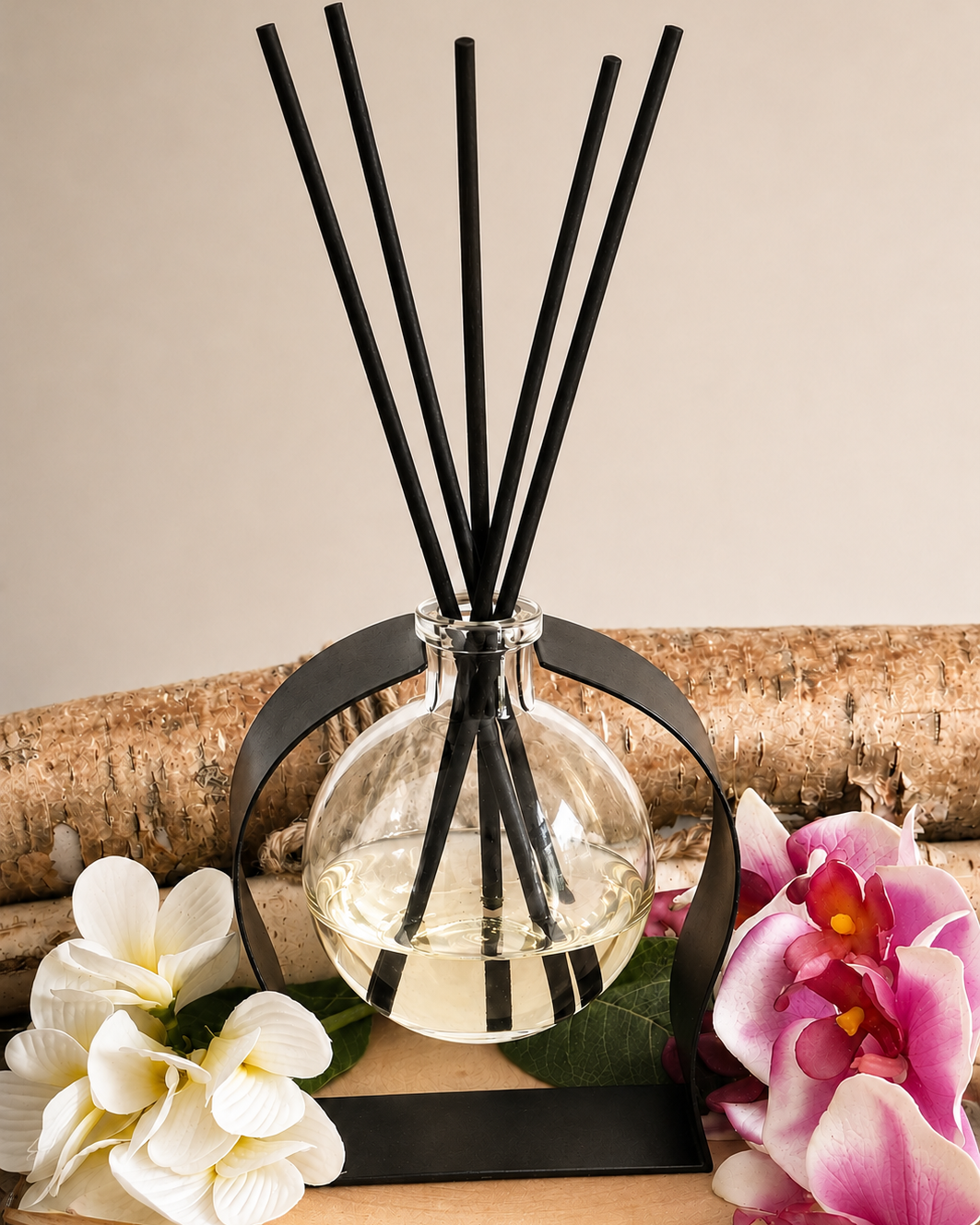 A glass reed diffuser bottle with clear liquid and black reeds, displayed on a wood slice with black metal bands, white flowers, and pink orchids against a beige background.