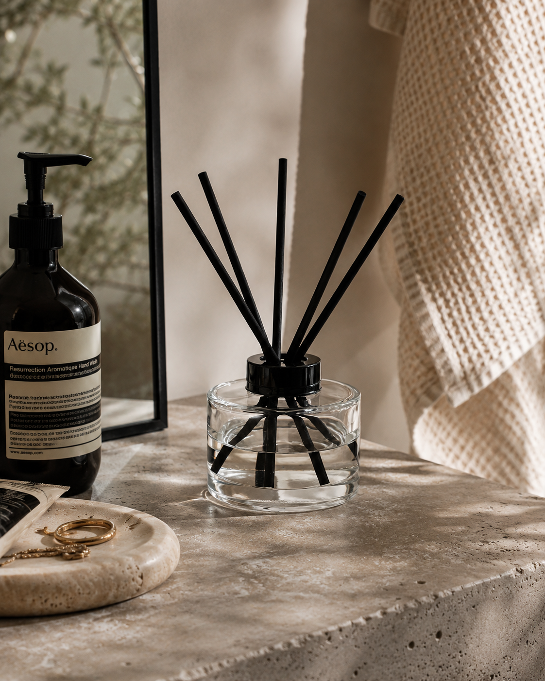 A clear glass reed diffuser with black reeds and a black cap sits on a stone surface beside a dark amber pump bottle and a textured cream towel in a softly lit interior setting.