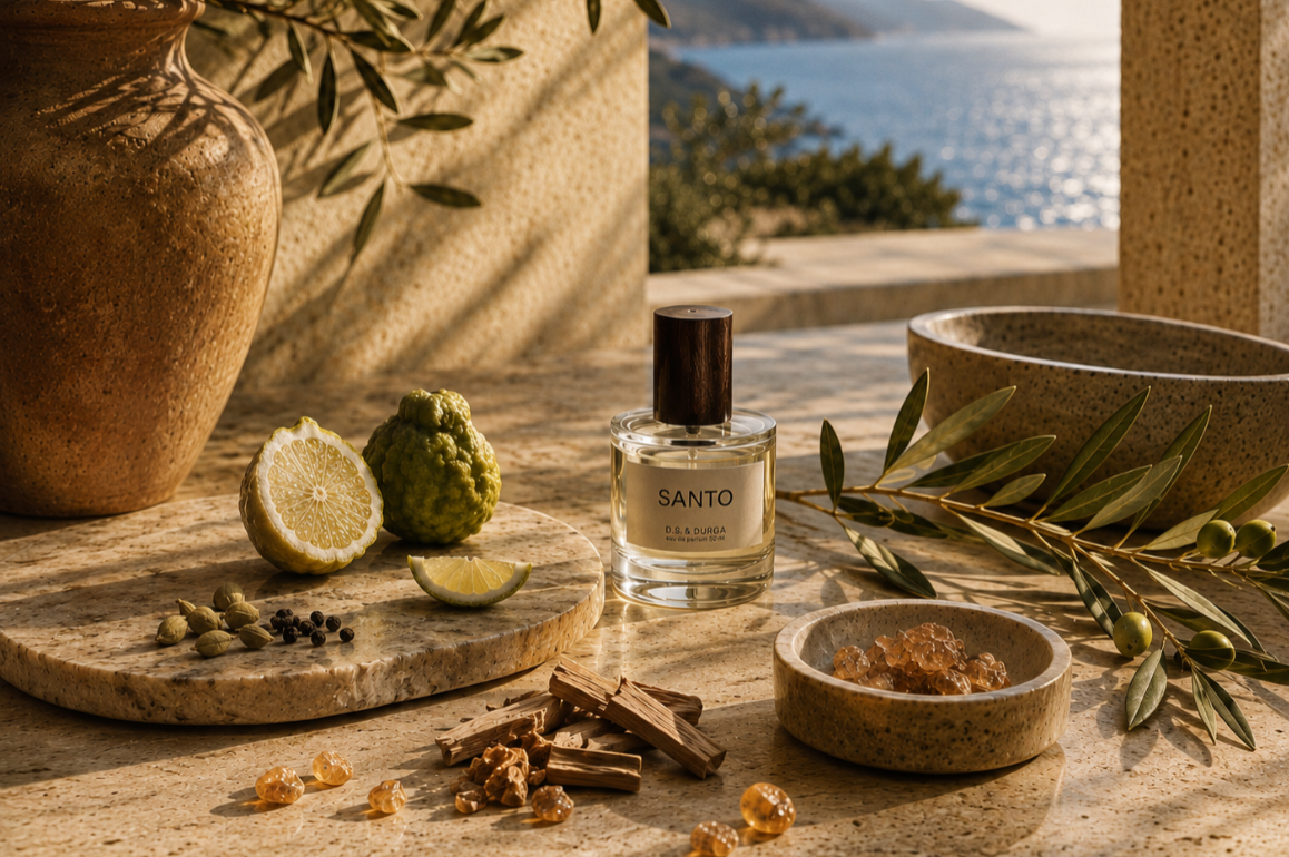 A clear glass perfume bottle labeled SANTO  with a black cap sits on a stone surface outdoors, surrounded by olive branches, a ceramic vase, citrus slices, spices, and a bowl. A coastal sea and mountains are visible in the background.