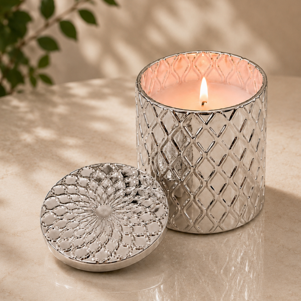 A silver textured candle jar with a matching lid sits on a light stone surface beside a lit candle. The scene is softly lit with leaf shadows, and a blurred plant is visible in the background.