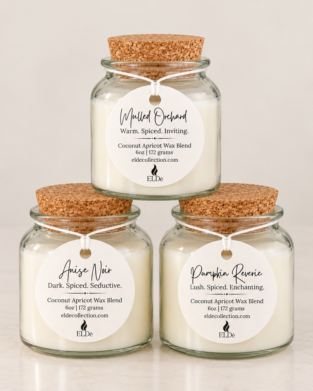 Three glass jar candles with cork stoppers are arranged in a pyramid on a light marble surface against a white background. The top candle is labeled 'Mulled Orchard' and the two bottom candles are labeled 'Anise Noir' and 'Pumpkin Revere'.