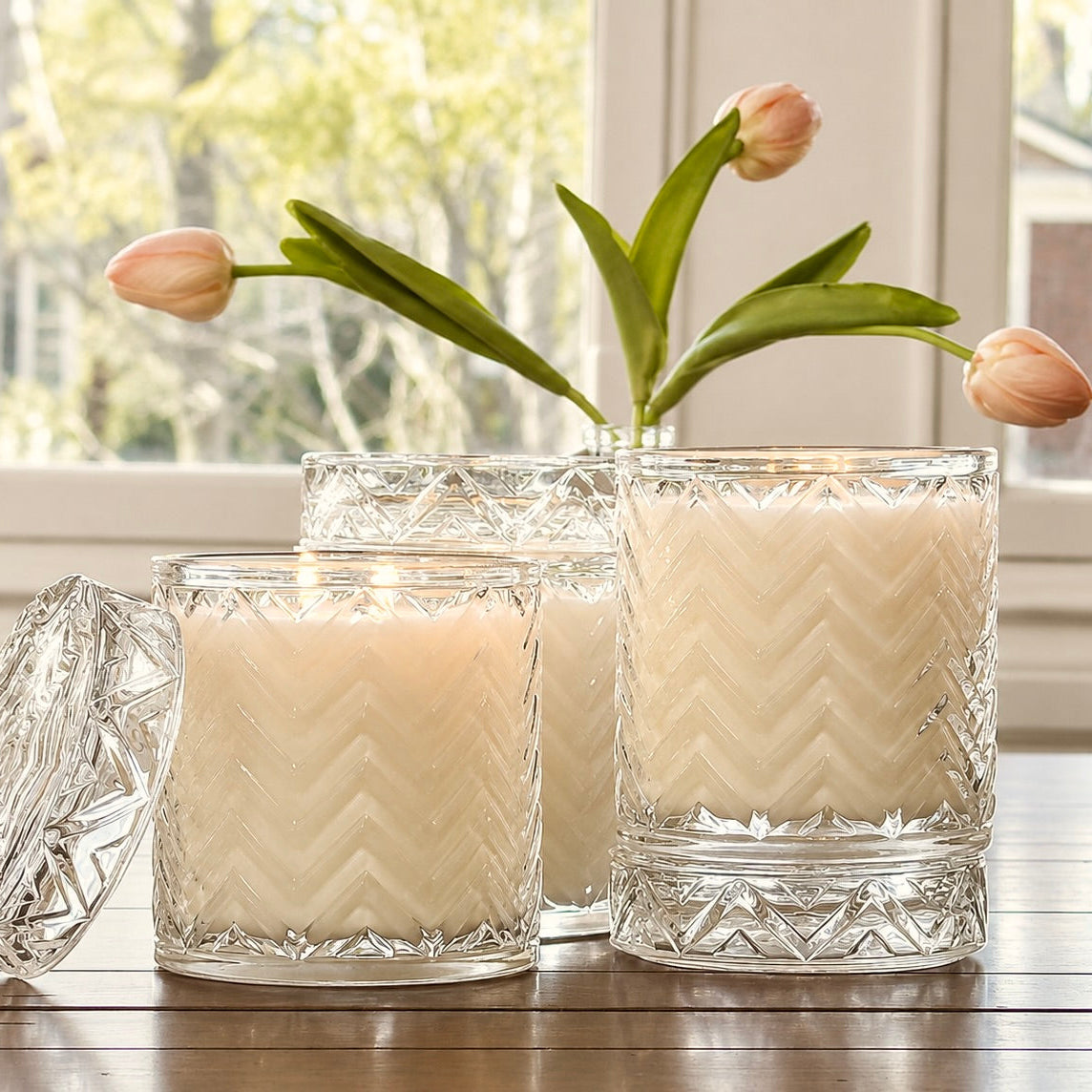 Three cream-colored candles in clear cut-glass jars with a chevron pattern, arranged on a wooden table in front of a window with tulips in the background.