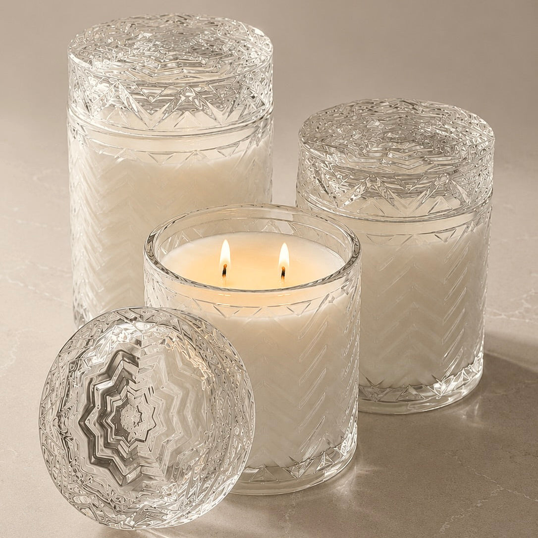 Three clear glass candle holders with textured, patterned lids and bodies, including one lit white candle in the center, set on a neutral beige surface.