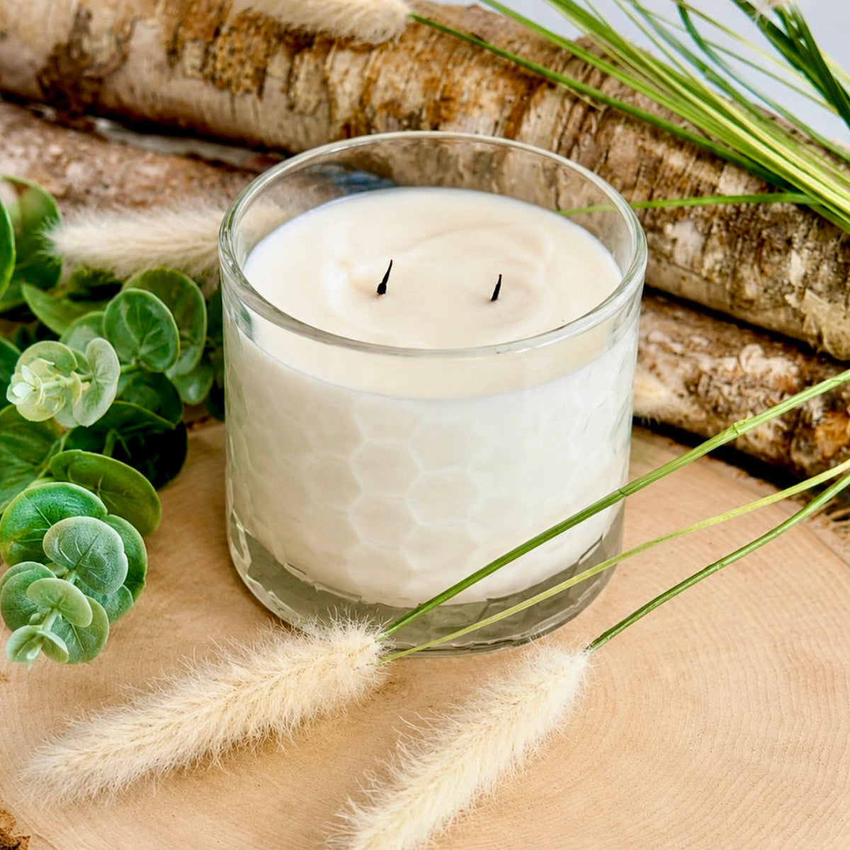 A white candle in a clear glass jar with a honeycomb-etched pattern, styled on a wooden slice with eucalyptus leaves, birch logs, and dried grasses.