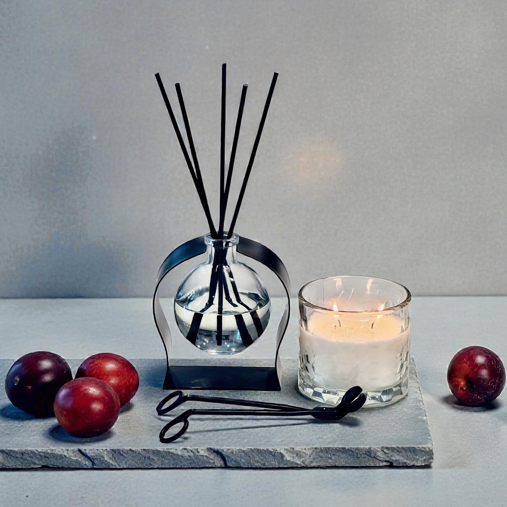 Aromatherapy diffuser with reeds, a candle, and plums on a gray surface.