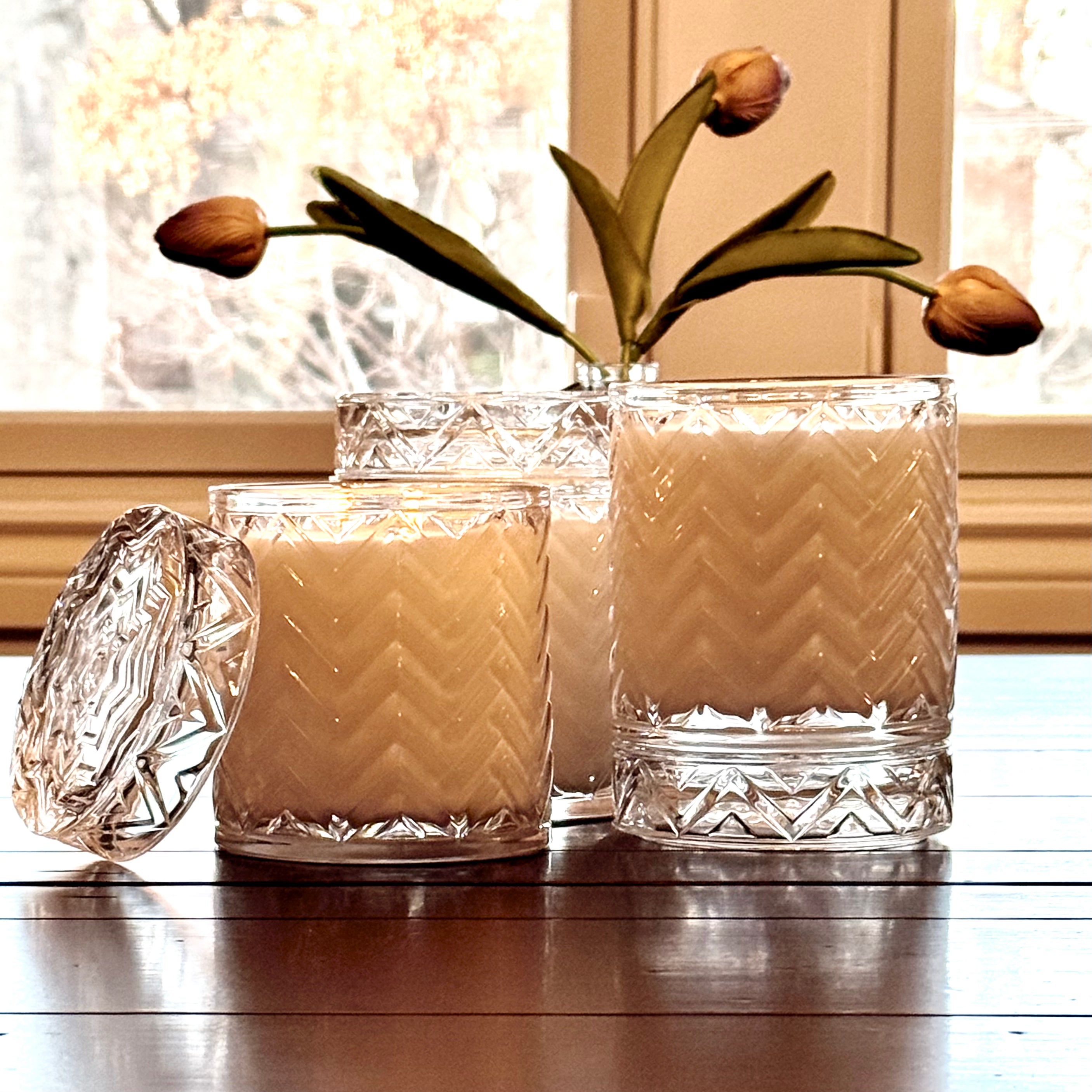 Two glass candles with decorative patterns on a wooden surface, with a blurred window in the background.