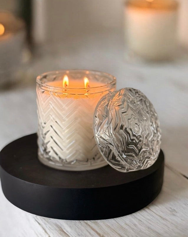Candle in a textured glass holder on a black coaster with a decorative lid, on a wooden surface.