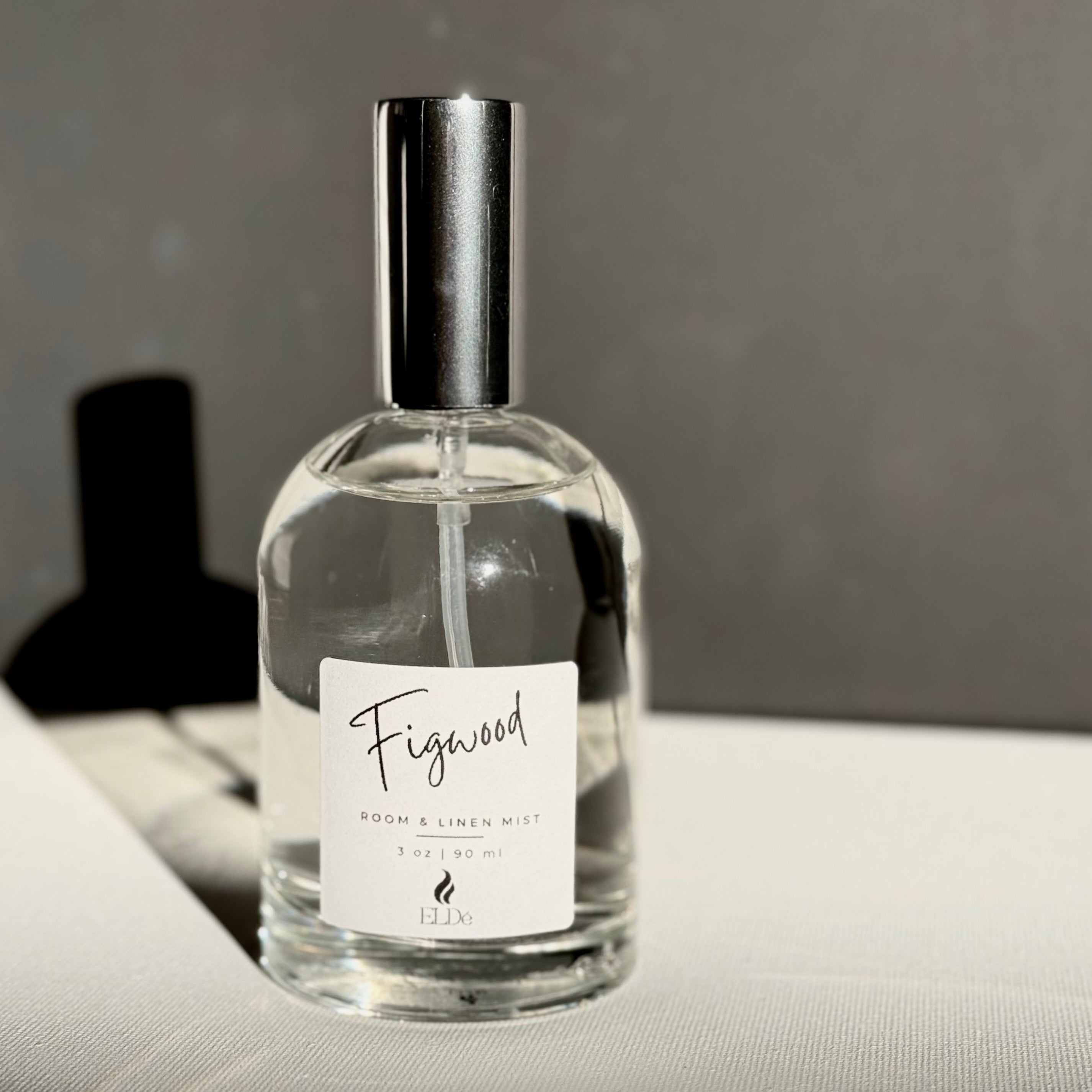 Clear bottle of Figwood room and linen mist on a textured surface with a blurred background