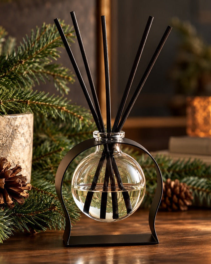 ELDé Collection Radiance diffuser in Frasier Fir, displayed with black reeds in a curved black stand, surrounded by fresh evergreen branches and pinecones in a cozy setting.
