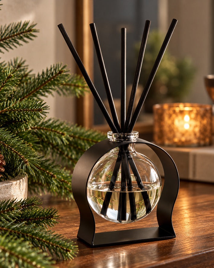 ELDé Collection Radiance reed diffuser in Frasier Fir, featuring a spherical glass bottle suspended in a black metal stand, styled with evergreen branches on a warm wooden table.