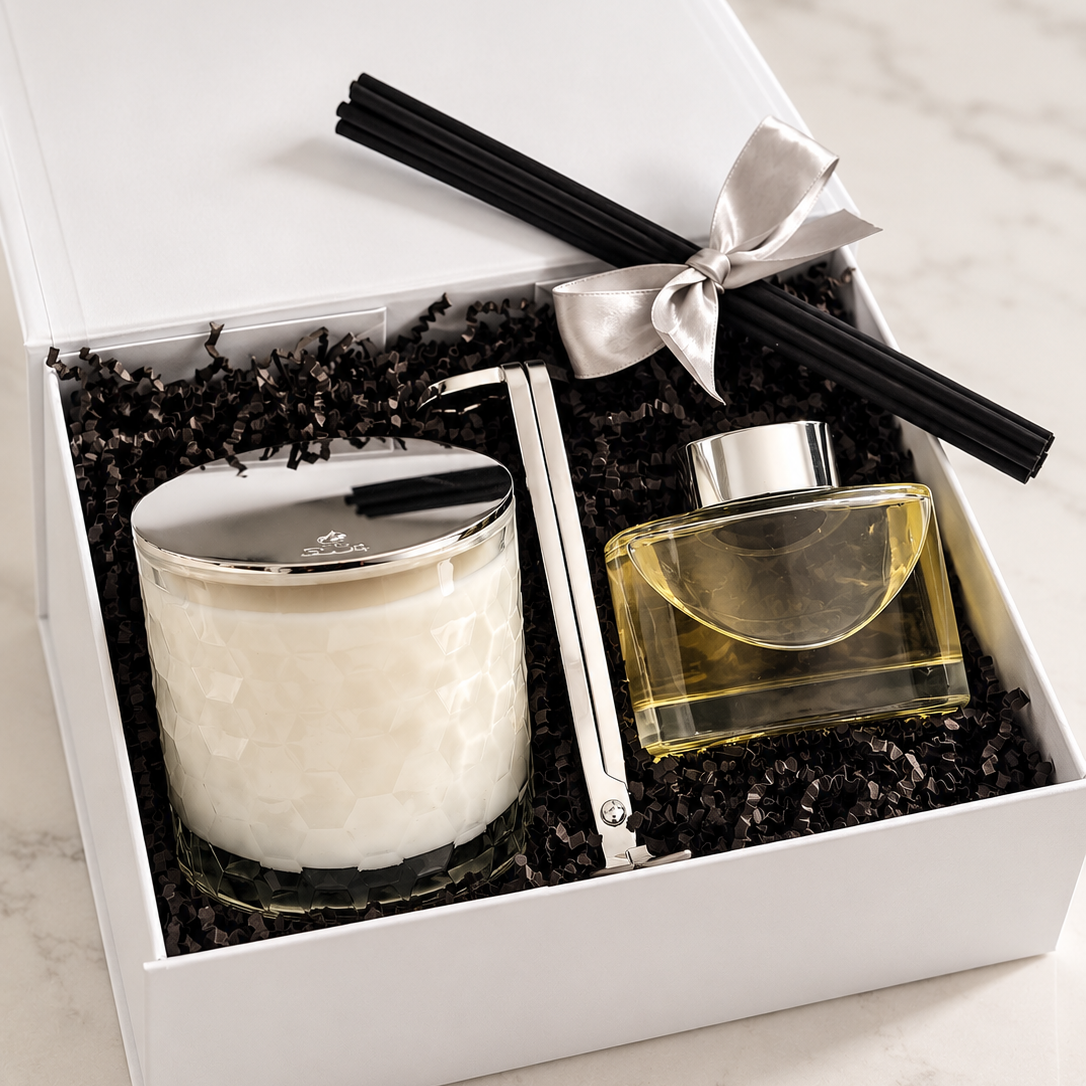 A gift set in a white box containing a white candle with a silver lid and a clear glass diffuser with liquid, and a silver wick trimmer all resting on black shredded paper. A silver ribbon bow and  black reed diffuser sticks are placed on top.