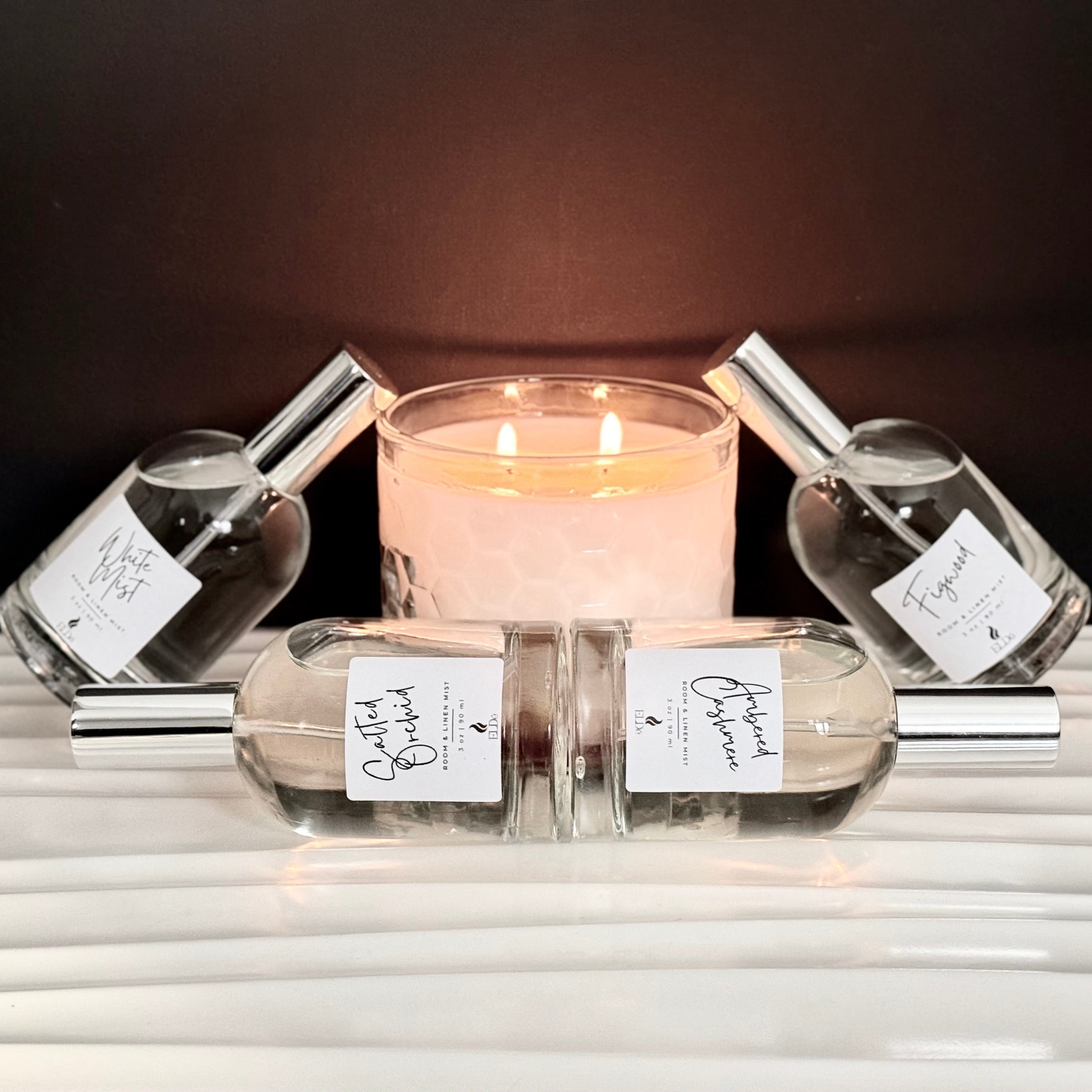 Candle surrounded by perfume bottles on a reflective surface with a dark background