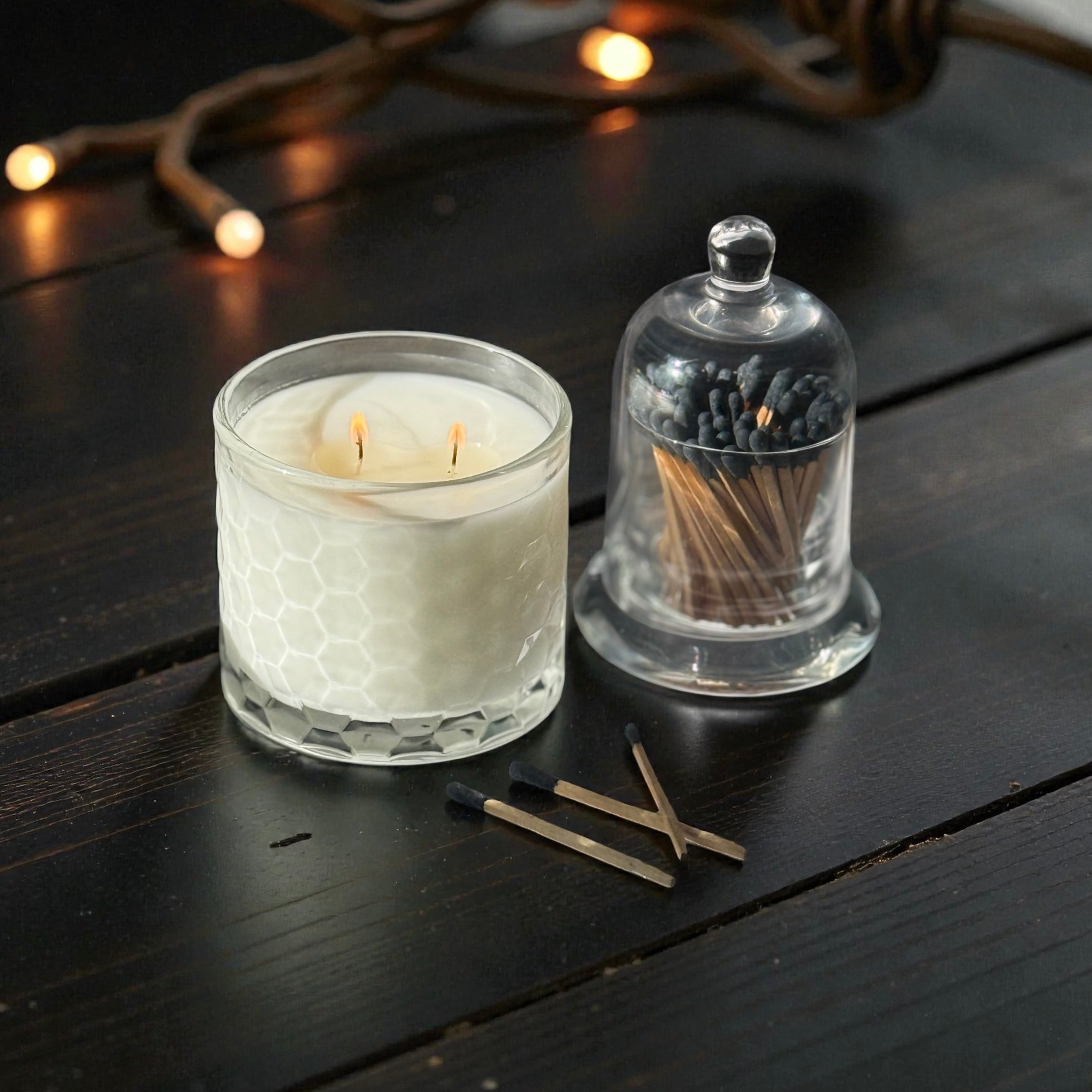White candle in a glass holder with a clear dome cover on a dark wooden surface.