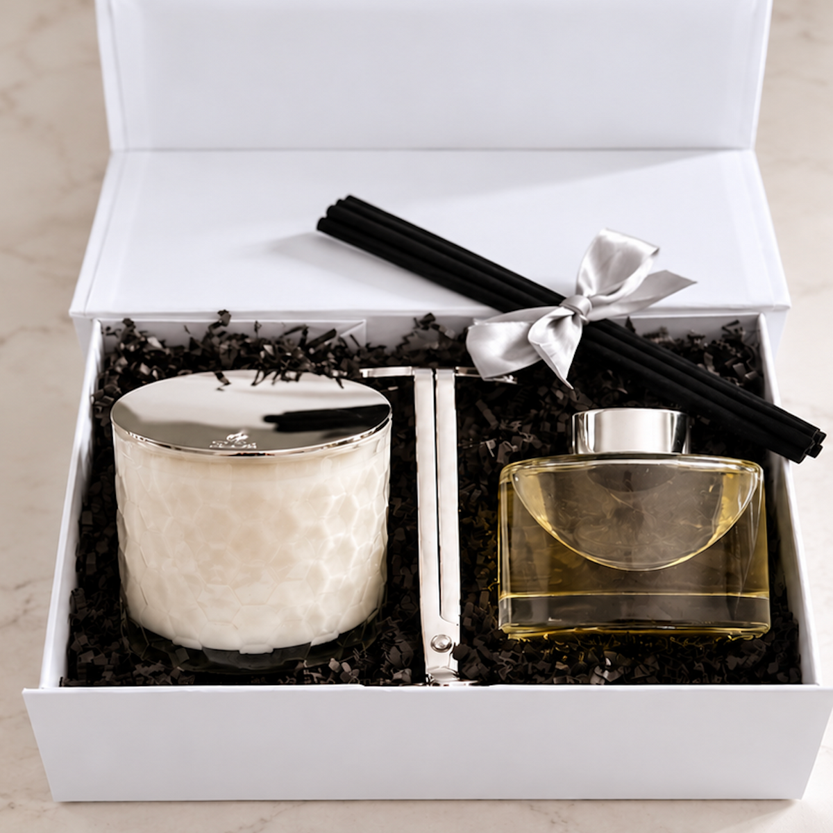 A luxury gift set in an open white box, featuring a white textured candle with a silver lid, a clear glass perfume bottle filled with golden-yellow liquid, and four black reed diffuser sticks tied with a silver ribbon.