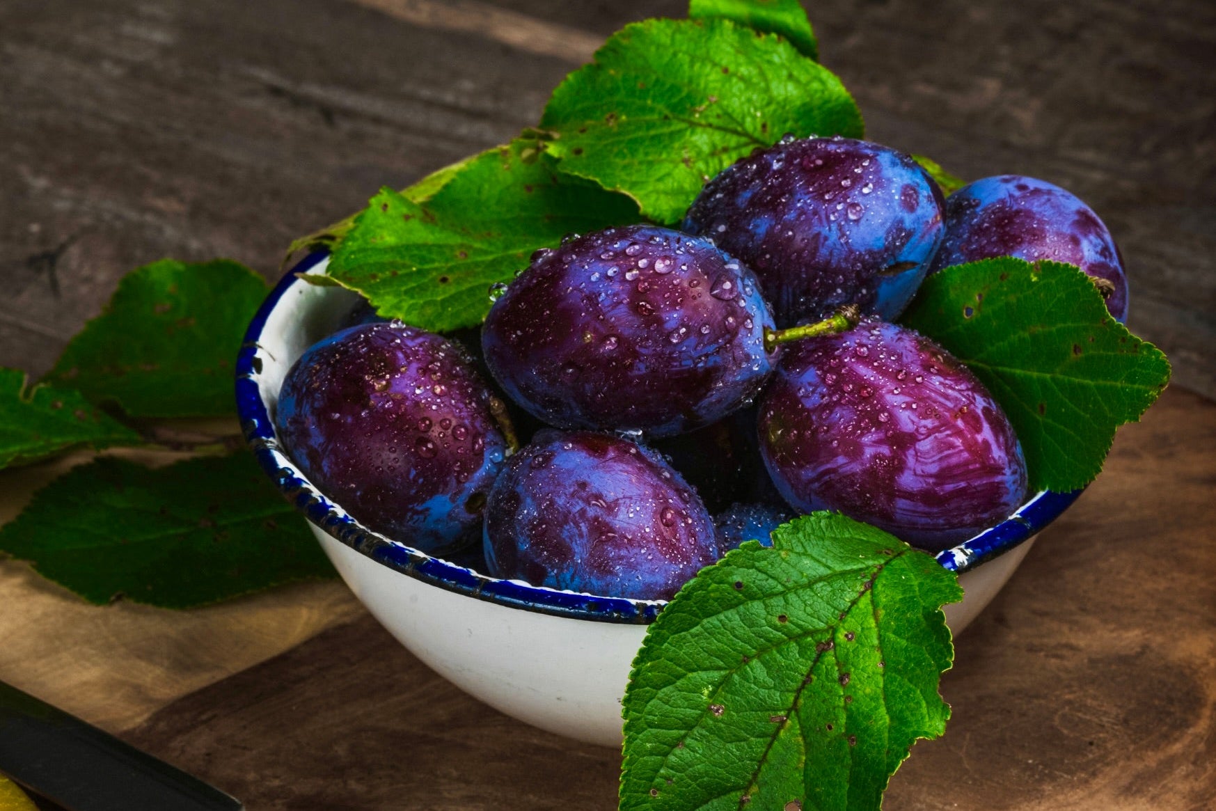 Bowl of plums with green leaves on a wooden surface