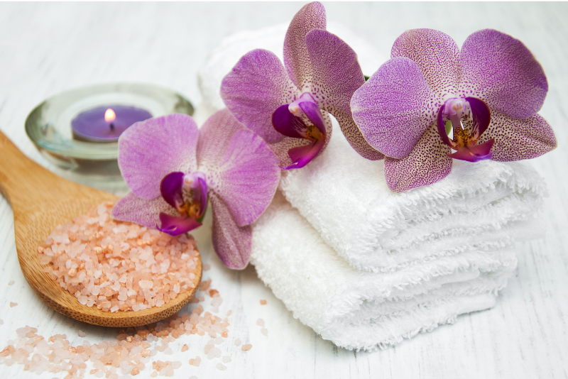 Purple orchids on a white towel with a wooden spoon of salt and a candle in the background.