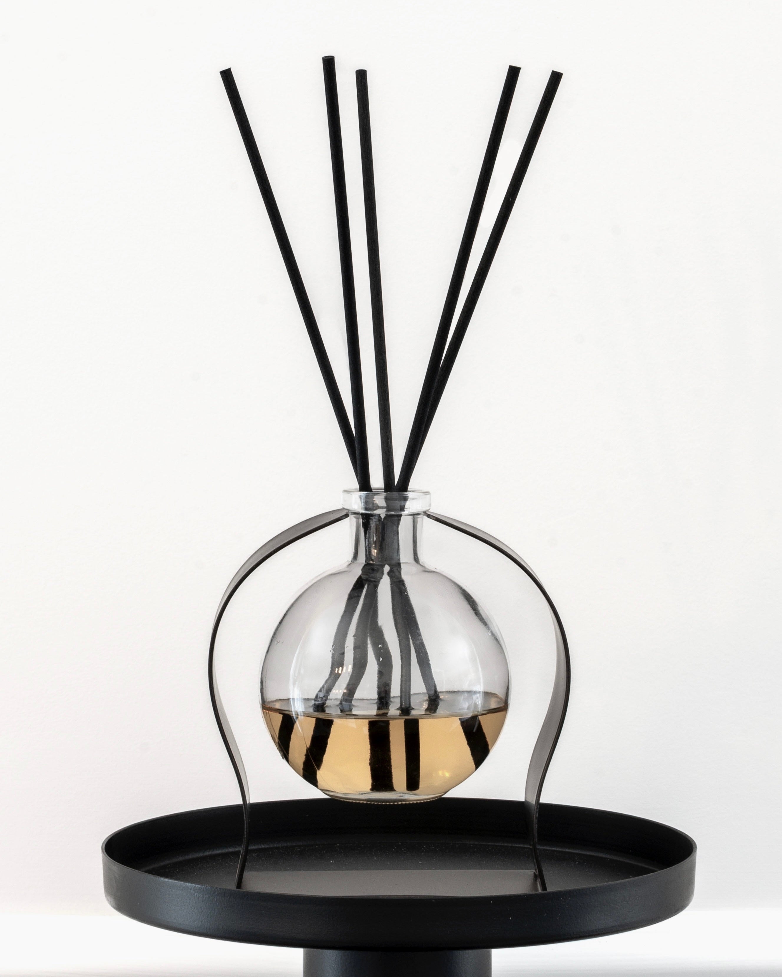 Decorative diffuser with black sticks in a glass container on a black stand against a white background