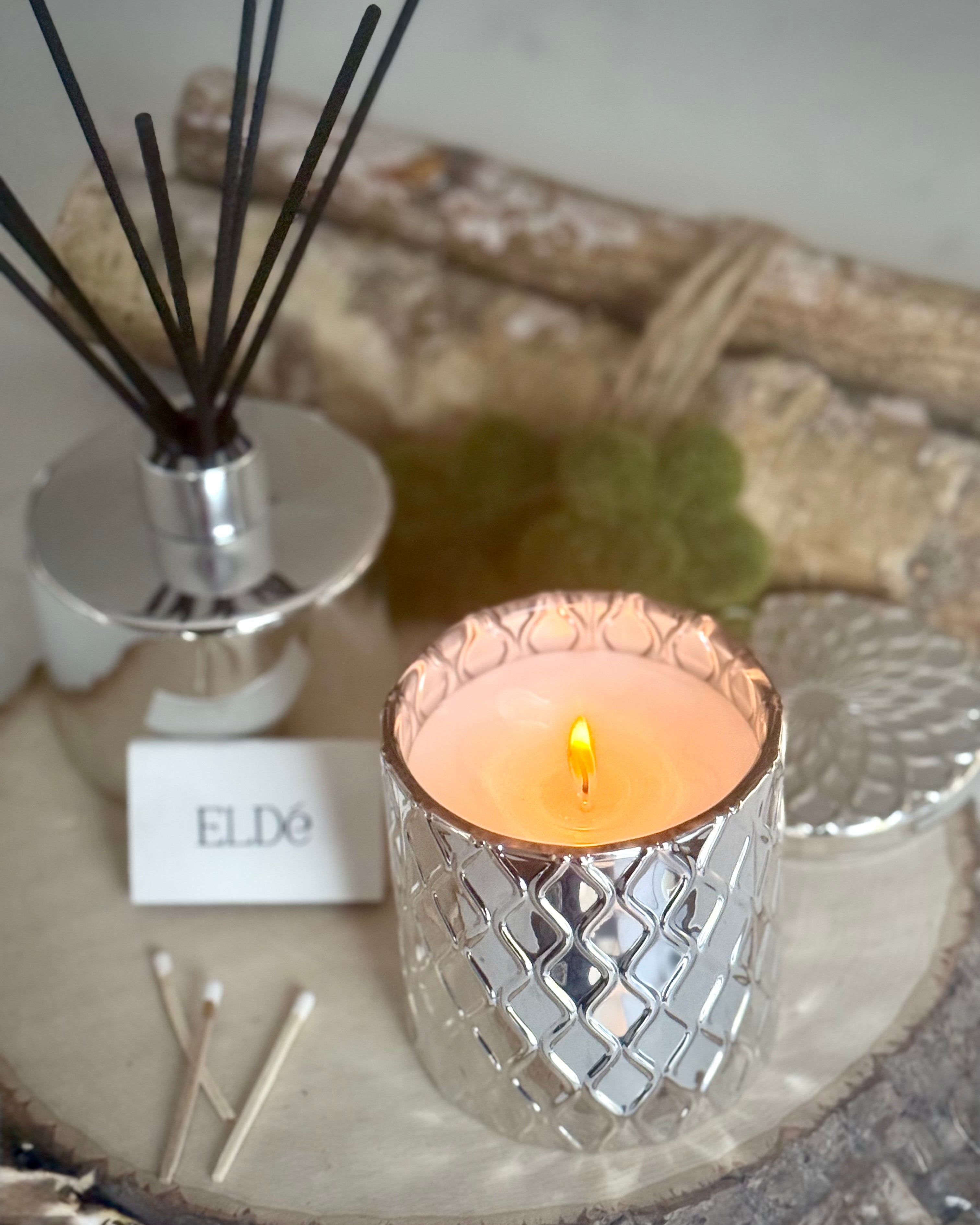 Candle in a decorative holder with reed diffuser and matches on a wooden surface