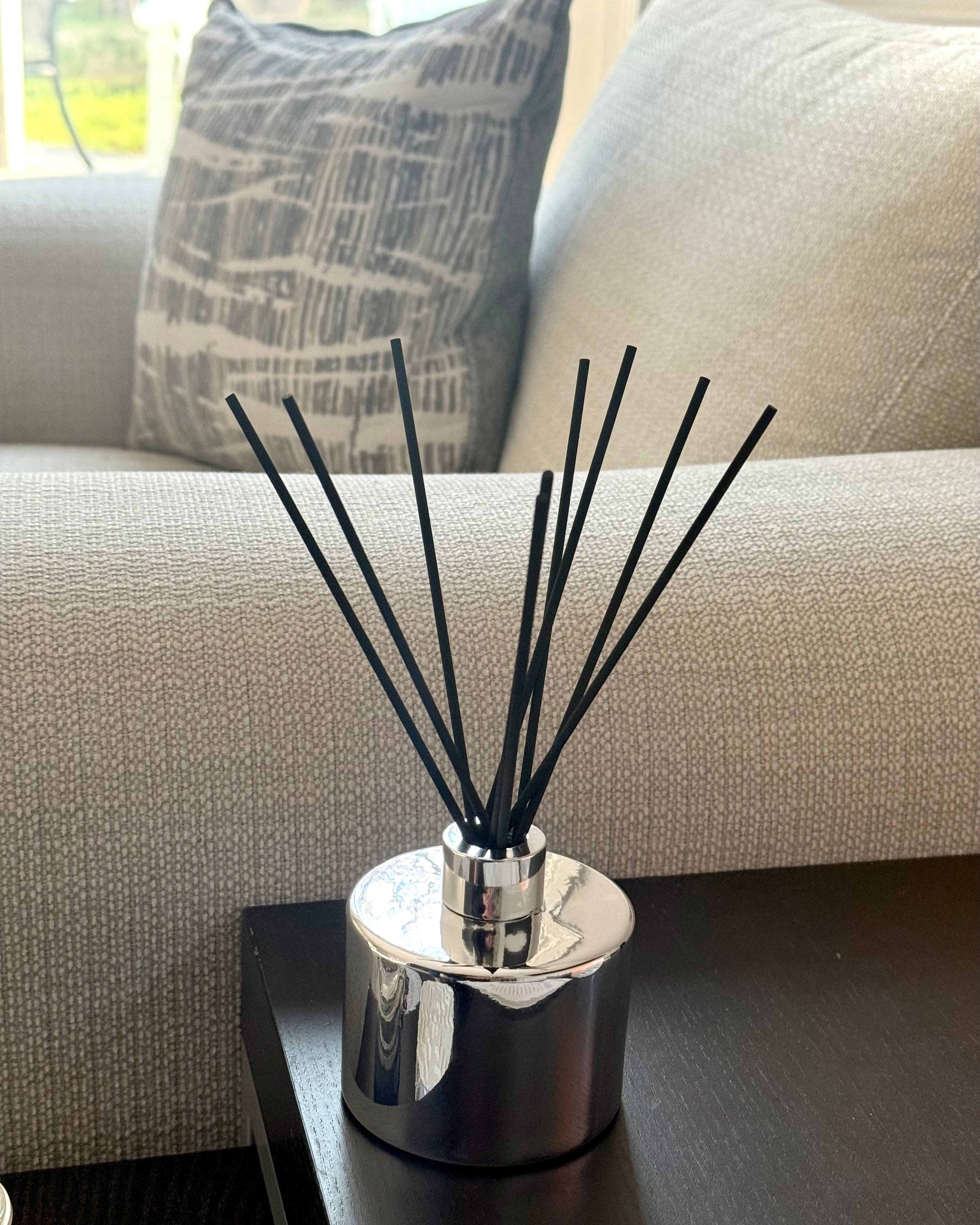 Black reed diffuser in a silver container on a coffee table with a couch and pillow in the background.