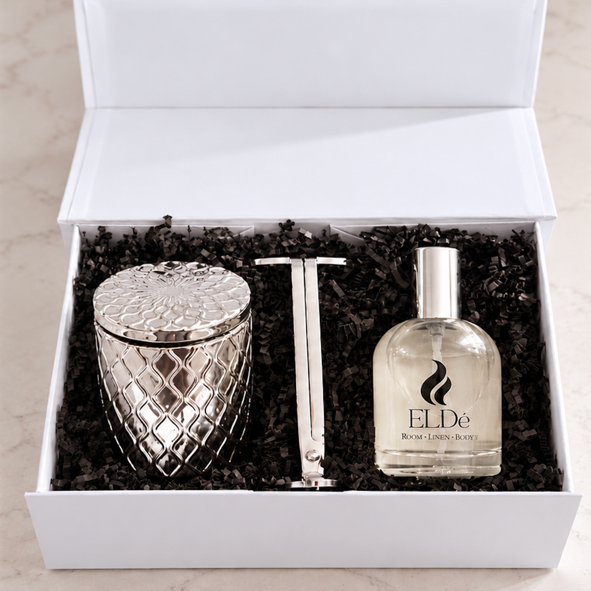 A gift set in an open white box with black shredded paper filler, containing a silver patterned metal container, a silver rectangular object, and a clear perfume bottle labeled ELDe Rose Parfum.