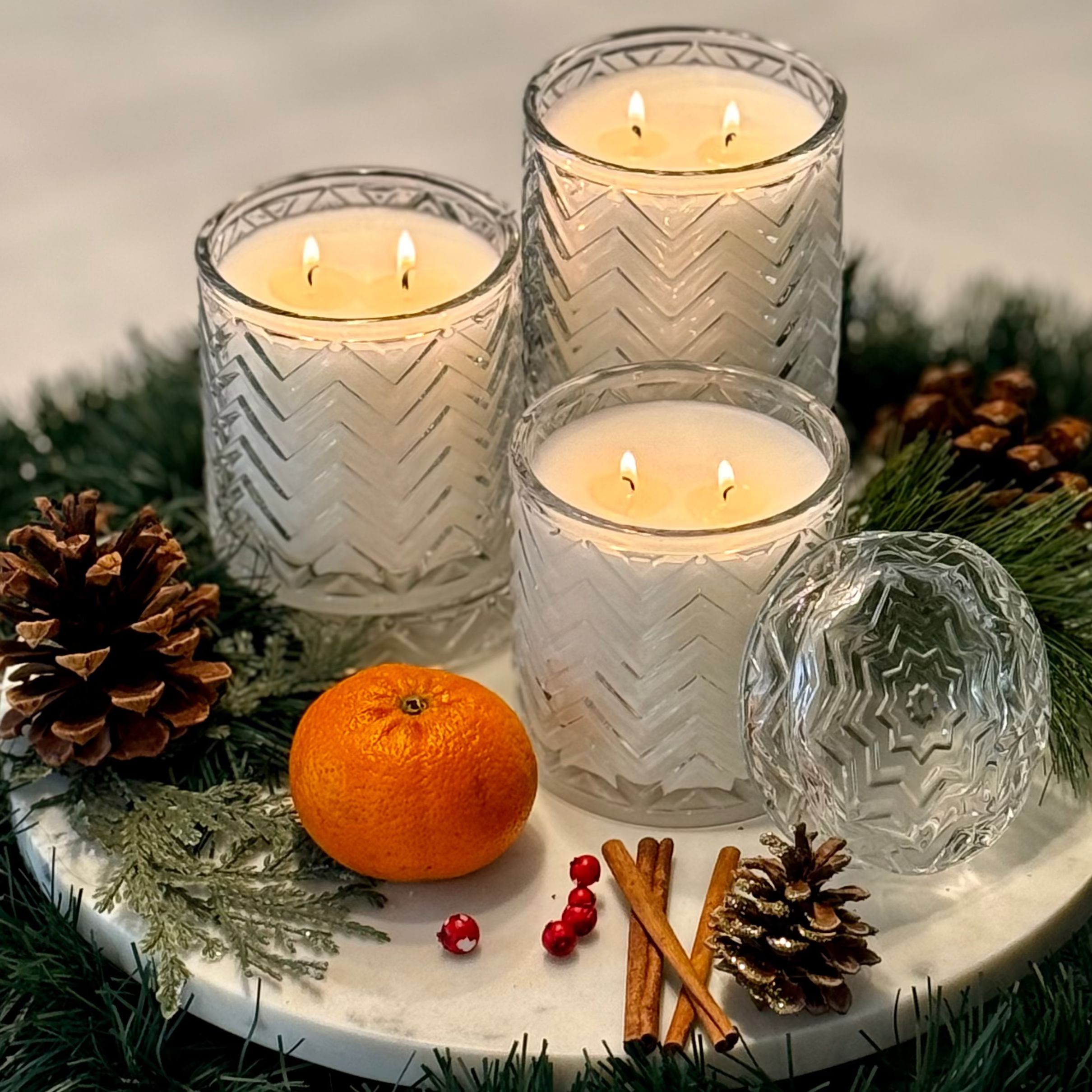 Four lit candles in textured glass holders on a decorative tray with an orange, cinnamon sticks, and pine cones.