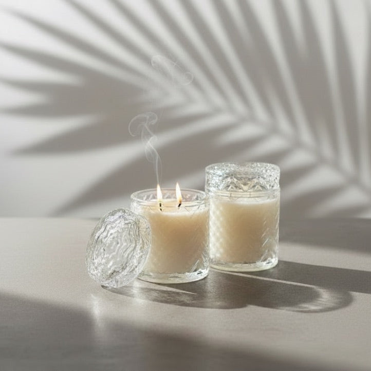 Two clear glass jar candles with cream-colored wax sit on a light surface, one lit with visible flames and smoke, against a white wall with palm leaf shadows.